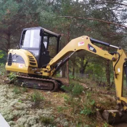 Żółta minikoparka CAT 302.5C na zalesionym terenie podczas wykonywania wykopu, widoczne ślady pracy i świeżo naruszona ziemia.