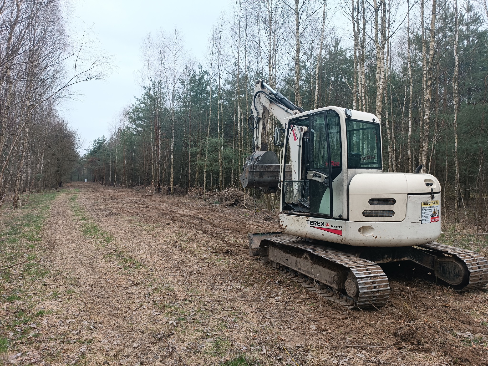 Biała koparka Terex Schaeff na leśnej drodze z wyciętymi drzewami po bokach, widoczne ślady opon i błoto, pochmurne niebo w tle.