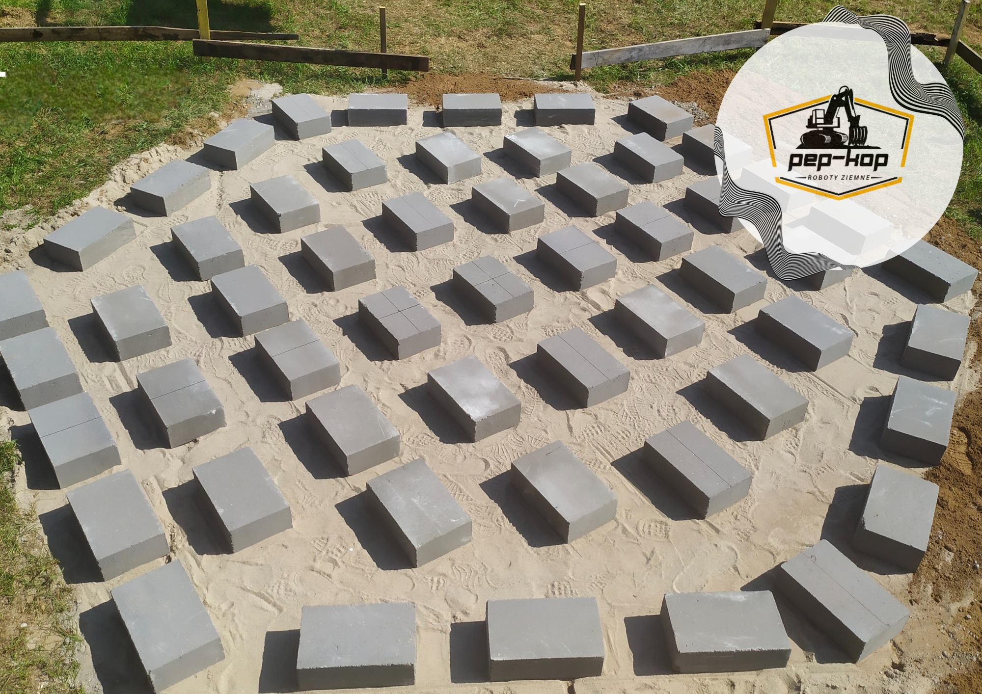 Przygotowany teren pod budowę z rozłożonymi bloczkami betonowymi, widoczny piasek, częściowo trawa i logo firmy w prawym górnym rogu.