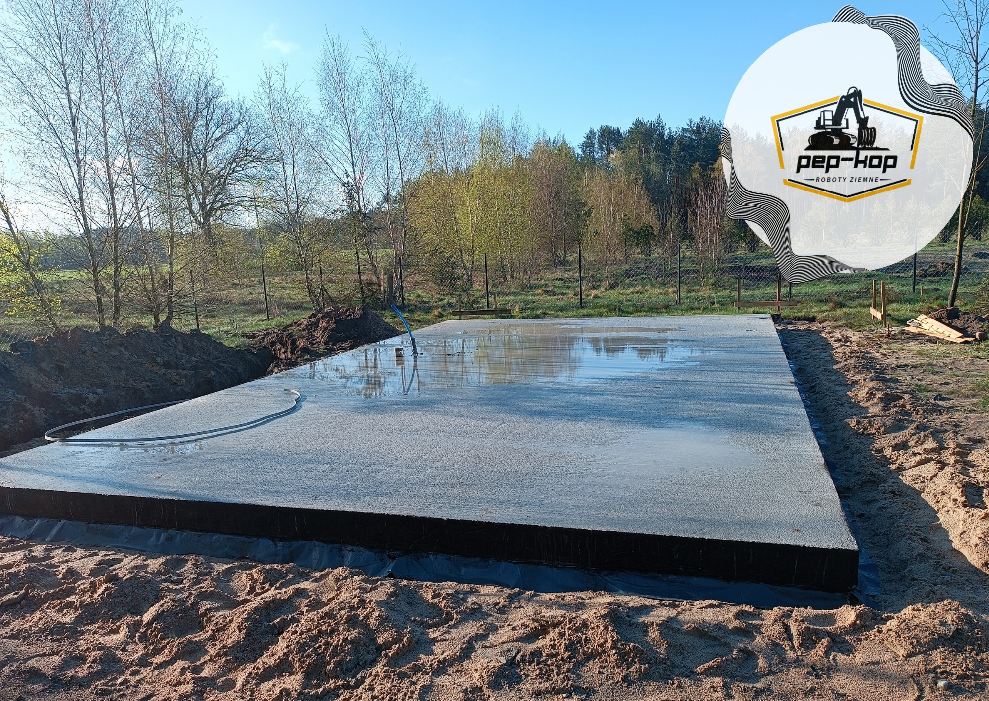 Świeżo wylana, mokra płyta fundamentowa na gruncie, zabezpieczona czarną folią, otoczona wykopem. W tle widoczne drzewa i logo firmy Pep-Kop Roboty Ziemne.