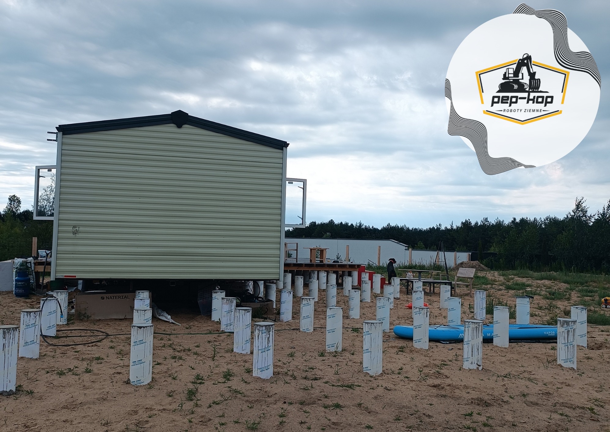 Montaż domu modułowego na przygotowanych, betonowych słupach fundamentowych z widocznym logo firmy Pep-Kop w prawym górnym rogu, na piaszczystym terenie pod pochmurnym niebem.