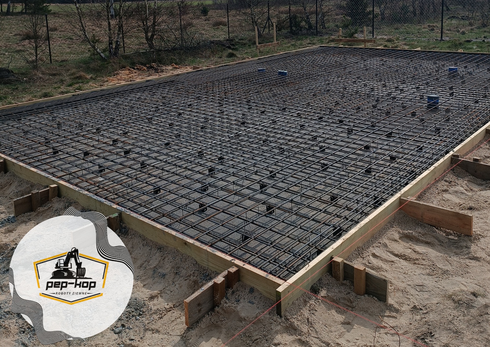 Zbrojenie płyty fundamentowej w drewnianym szalunku, widoczne podkładki dystansowe i logo firmy w rogu zdjęcia.