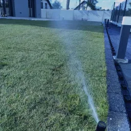 Zraszacz trawnikowy w akcji, nawadniający świeżo założony trawnik przy nowoczesnym domu z szarą elewacją i betonowym ogrodzeniem, widoczne krople wody w powietrzu.