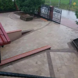 Ułożona kostka brukowa na podjeździe, z widocznym wzorem i obramowaniem z ciemniejszej kostki, brama wjazdowa z kutego żelaza w tle, widok z góry.