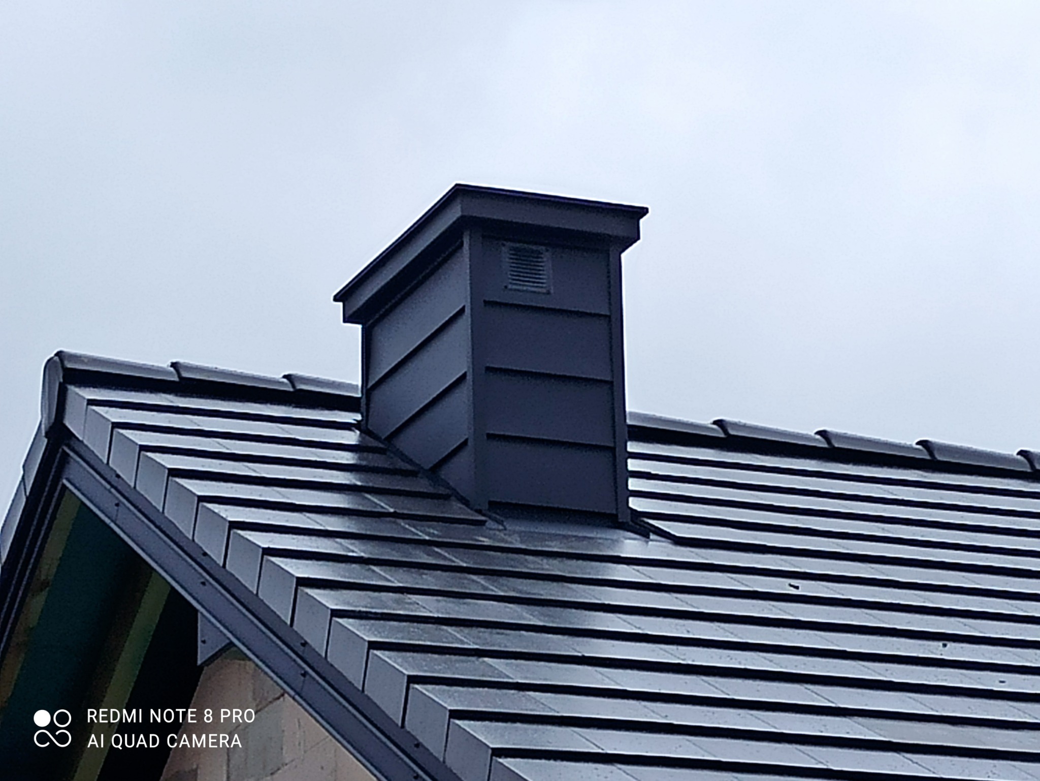 Błyszczący, grafitowy dach z dachówki ceramicznej z kominem wentylacyjnym w kształcie prostopadłościanu, sfotografowany pod kątem w pochmurny dzień.