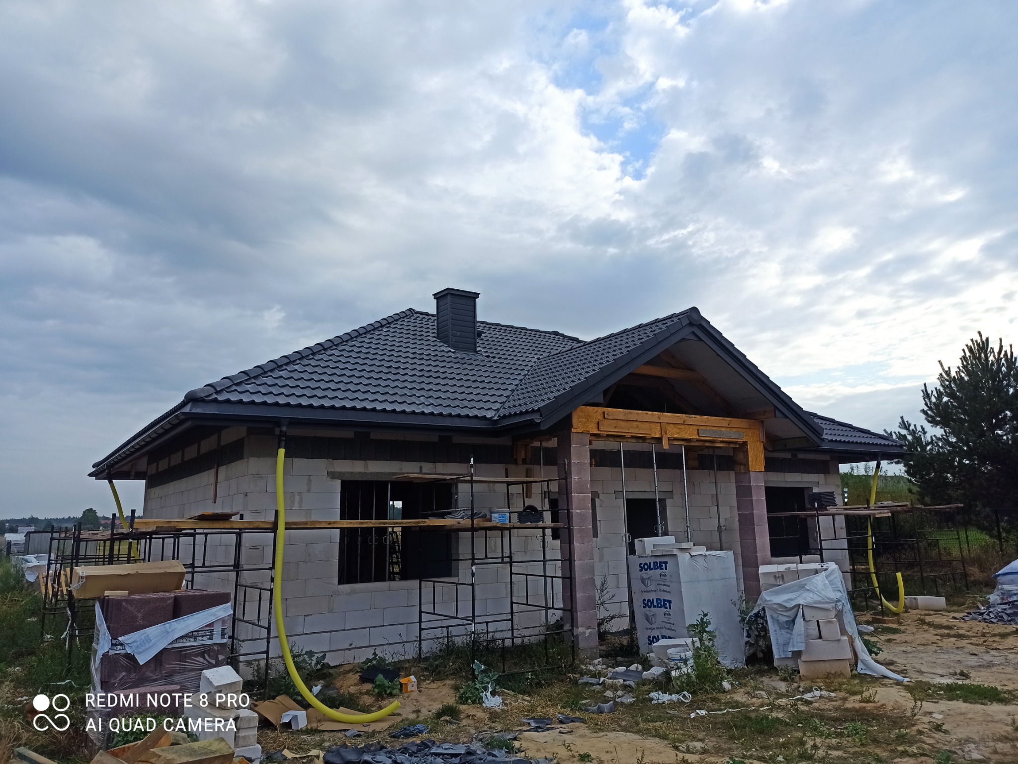 Dom w trakcie budowy z widoczną konstrukcją dachu pokrytą czarną dachówką, z kominem i fragmentem więźby dachowej, materiały budowlane Solbet na pierwszym planie.