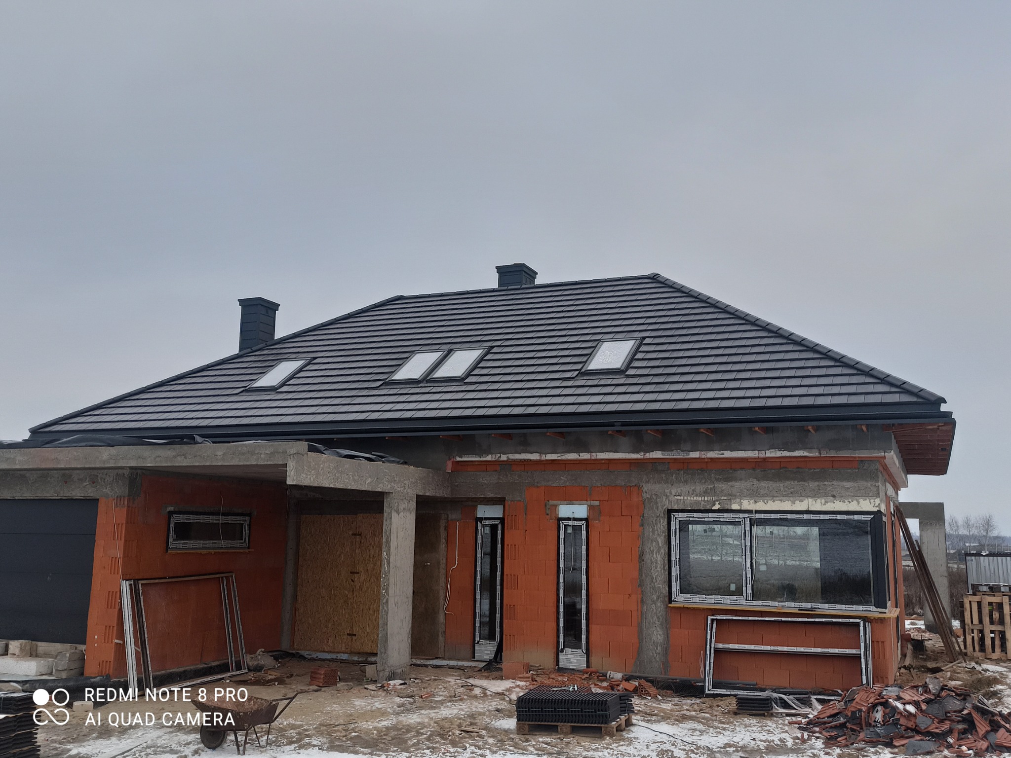 Dom jednorodzinny w trakcie budowy, widoczny dach z czarną dachówką z trzema oknami dachowymi, komin, surowy stan ścian z czerwonej cegły, okna bez szyb, teren budowy.