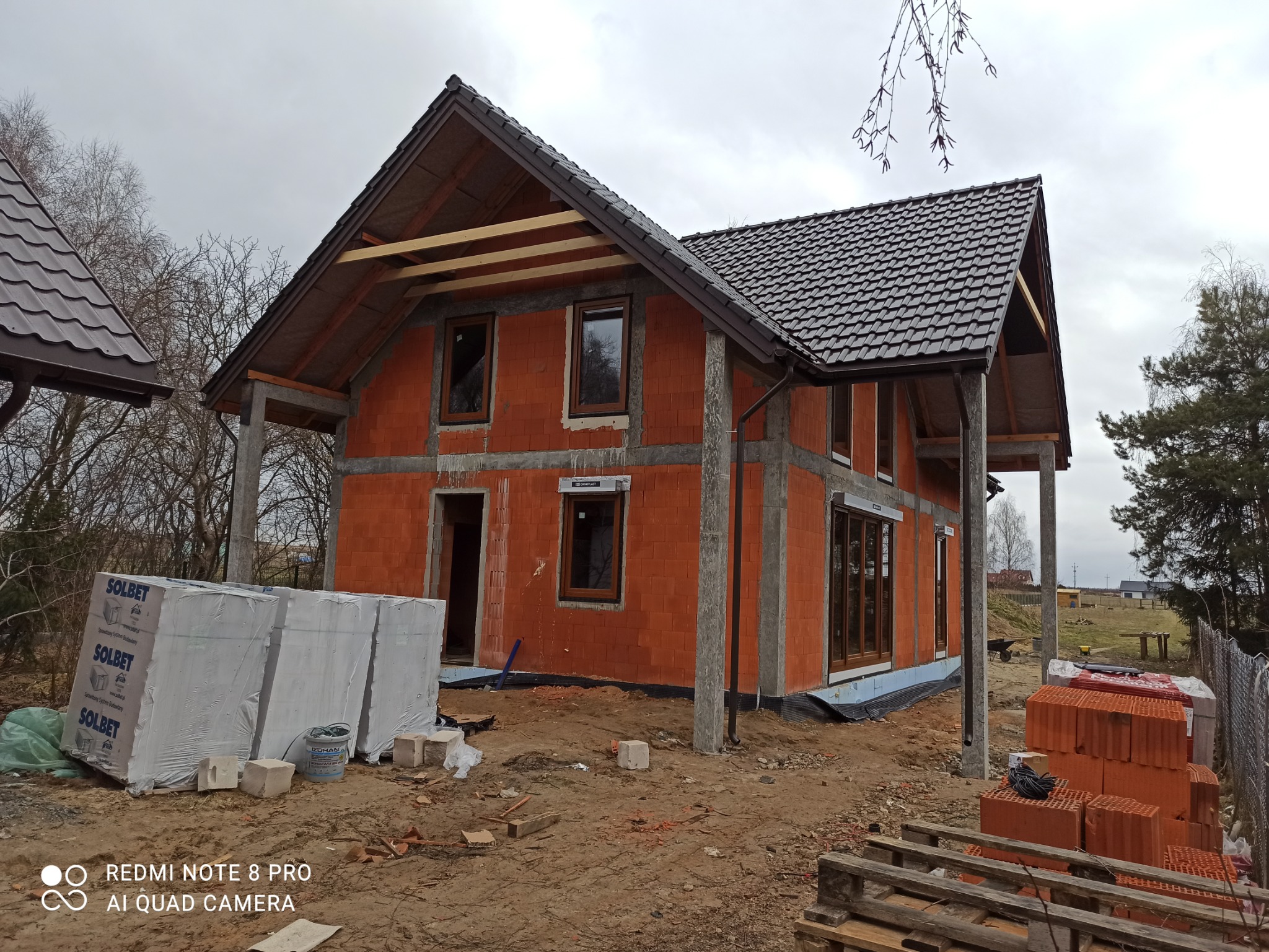 Surowy stan domu jednorodzinnego w trakcie budowy, widoczny dach z ciemnej dachówki, ściany z czerwonej cegły, okna z brązowymi ramami oraz materiały budowlane składowane na posesji.