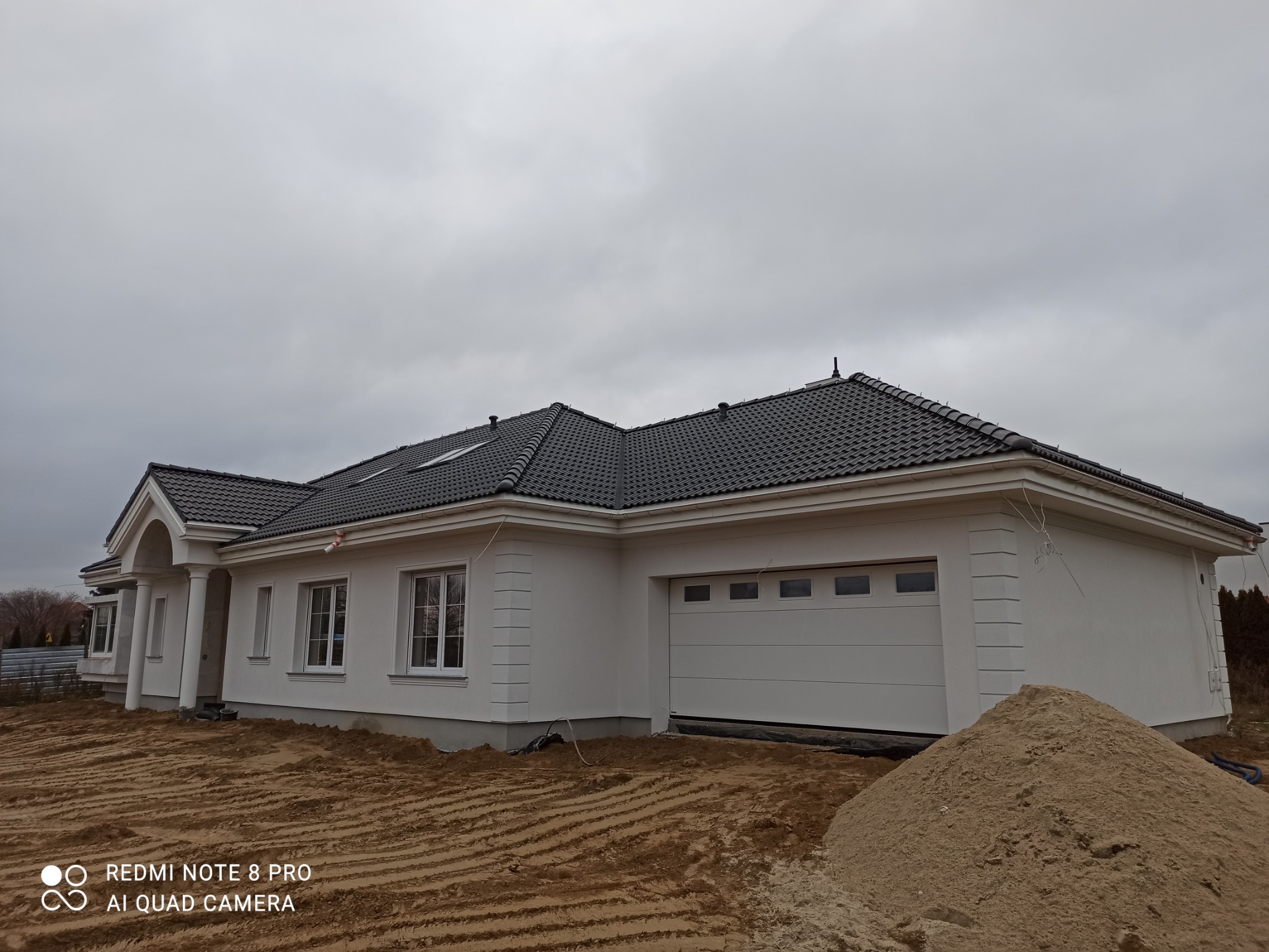 Nowoczesny parterowy dom jednorodzinny z ciemną dachówką ceramiczną i jasną elewacją, widok z zewnątrz na tle pochmurnego nieba i z pryzmą piasku na pierwszym planie.