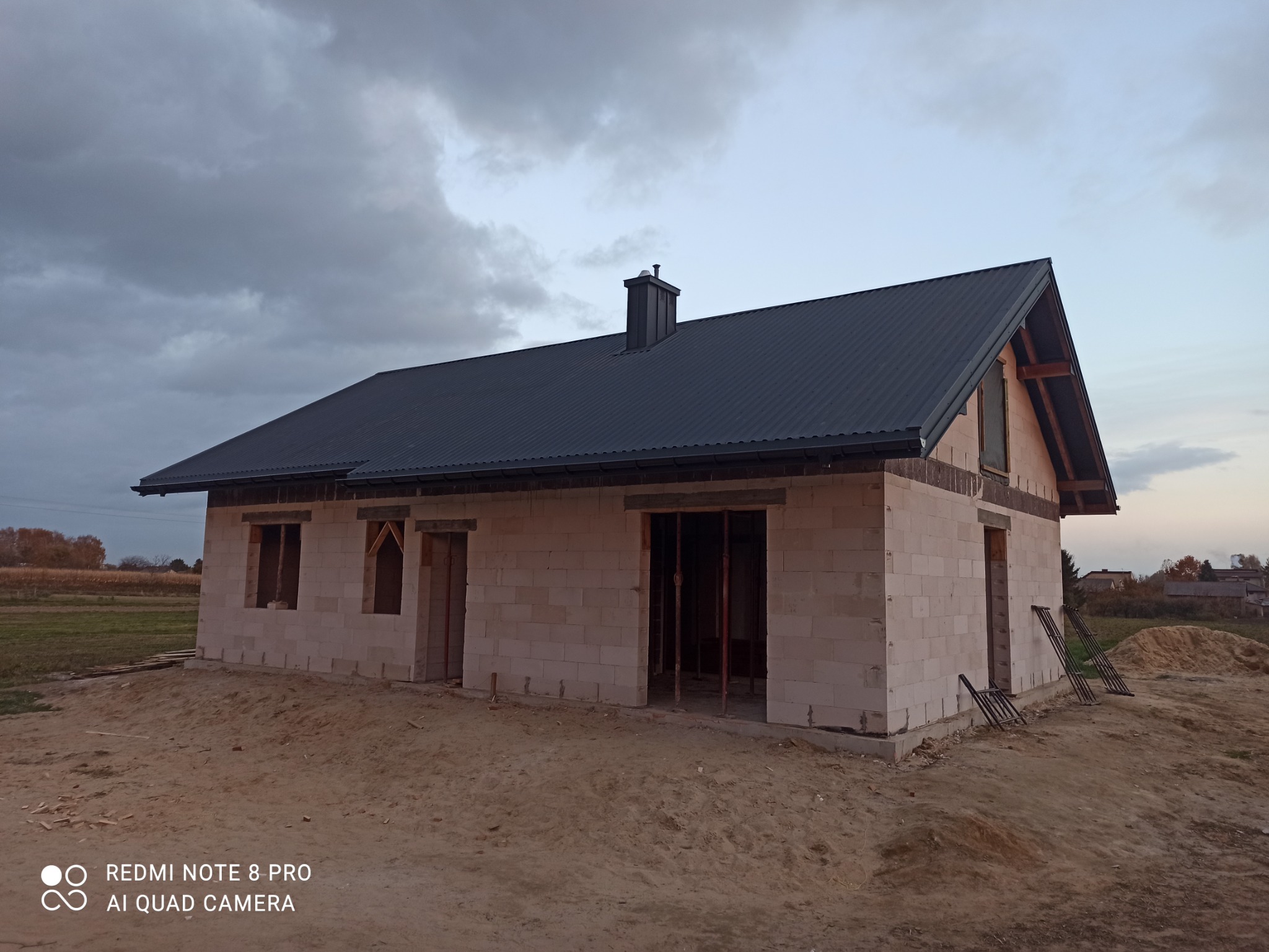 Dom w stanie surowym zamkniętym z ciemnym, metalowym dachem i kominem, widok z zewnątrz na tle pochmurnego nieba.