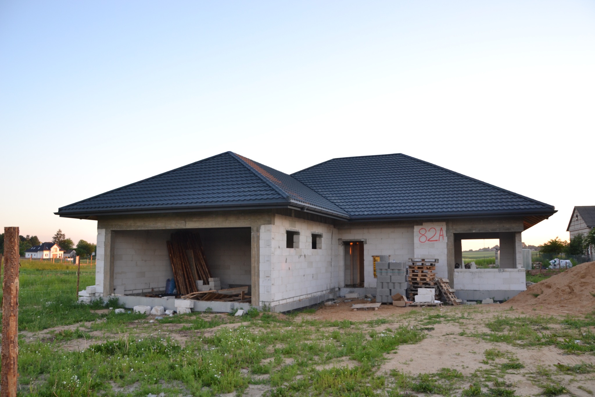 Nowo wybudowany dom jednorodzinny z ciemnoszarą dachówką, w stanie surowym otwartym, z widoczną stertą desek w garażu i numerem 82A na ścianie.