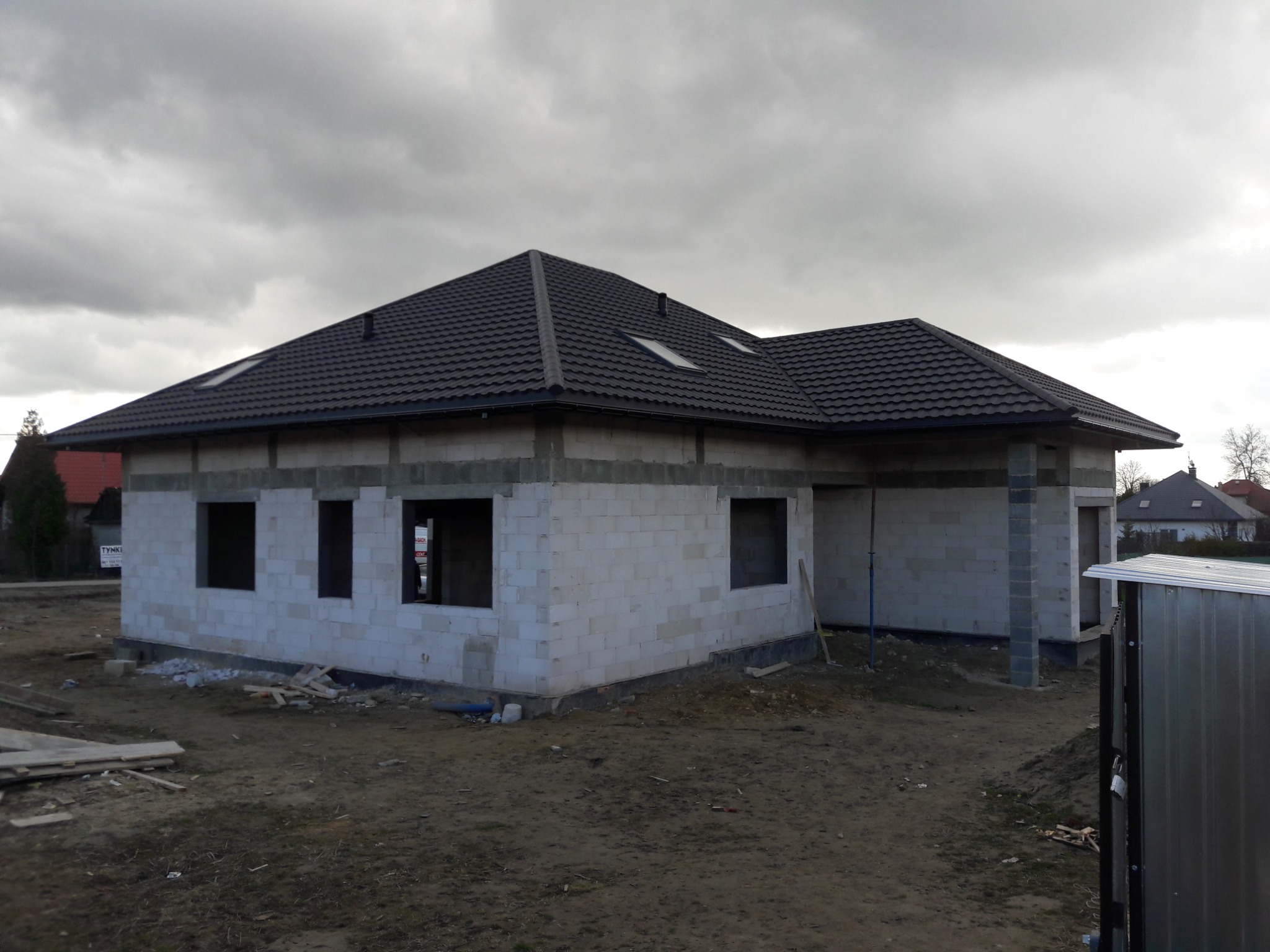 Dom jednorodzinny w trakcie budowy, z widocznym czarnym dachem z dachówki ceramicznej, oknami dachowymi i nieotynkowanymi ścianami z białych bloczków, na tle pochmurnego nieba.