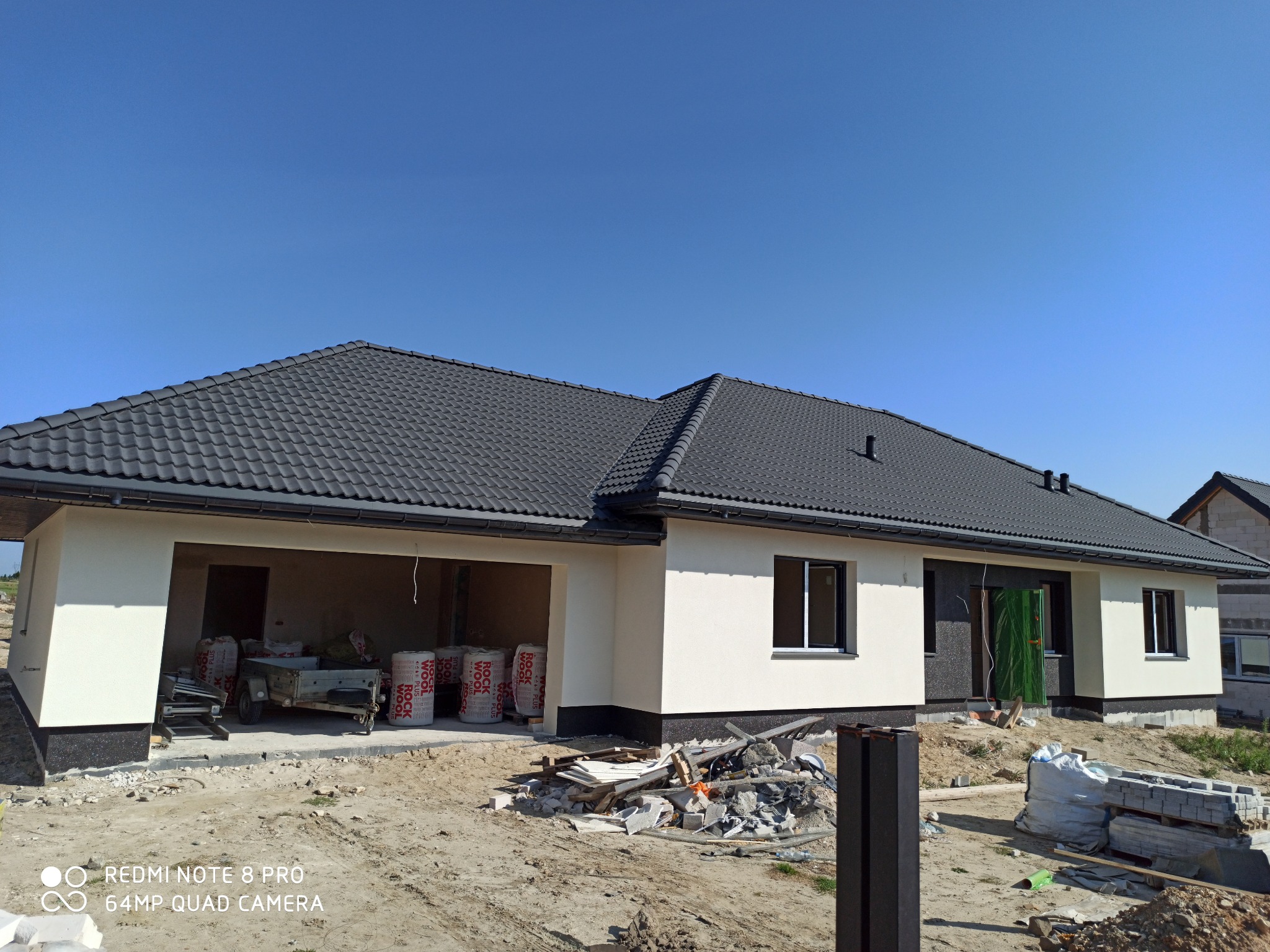 Nowoczesny dom jednorodzinny z ciemnoszarą dachówką ceramiczną, widoczne materiały budowlane i przyczepka w garażu, błękitne niebo w tle.