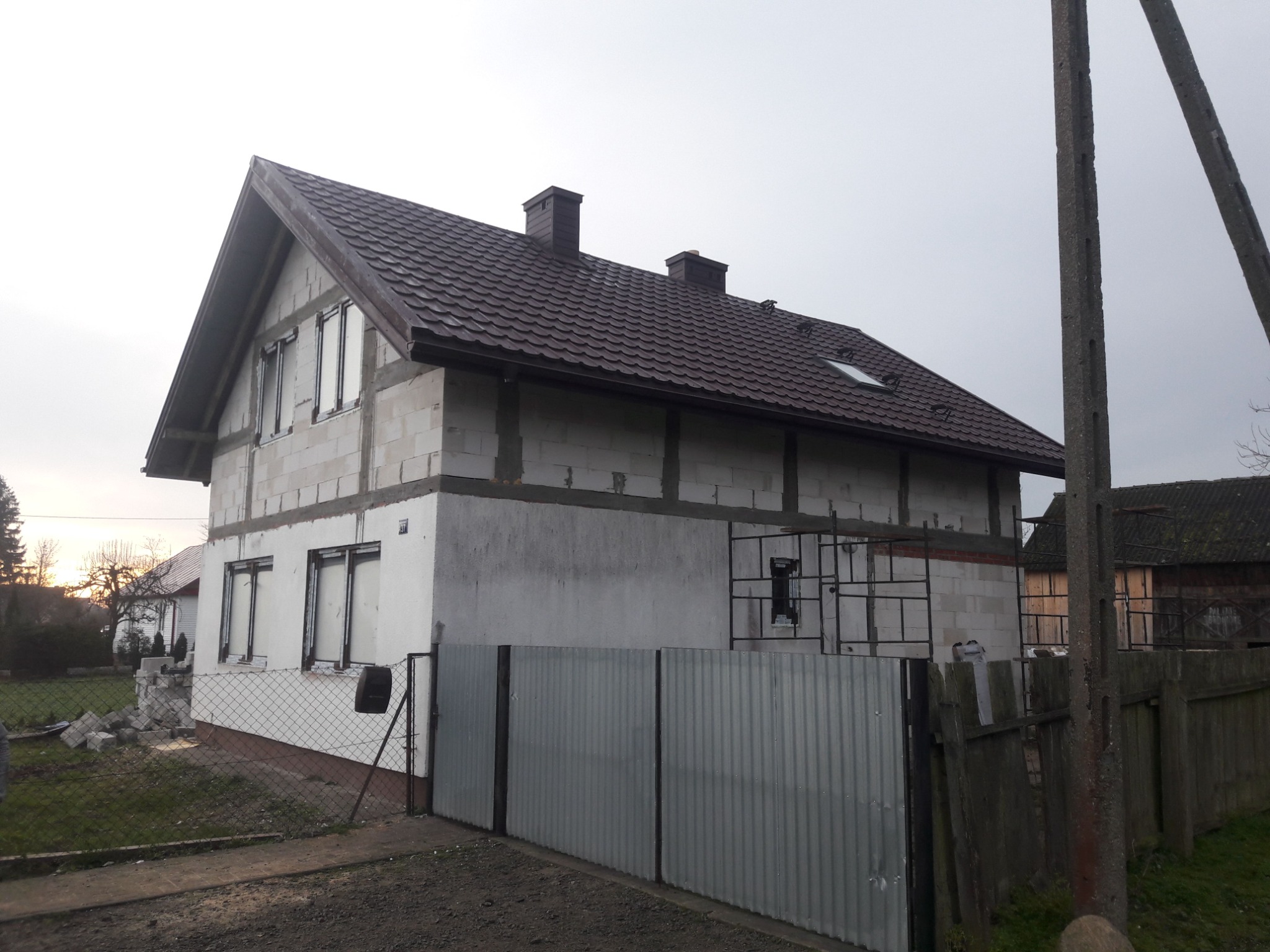 Dom w trakcie budowy z nowym, brązowym dachem z dachówki, widoczne kominy i okno dachowe, ściany w surowym stanie, częściowo otynkowane, szare ogrodzenie z blachy falistej.