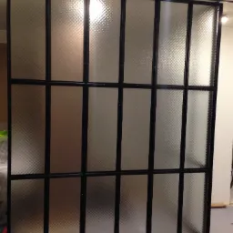 Metalowa konstrukcja ścianki działowej z wypełnieniem ze szkła ornamentowego, czarne profile, widoczne mocowania śrubowe, surowy stan wykończenia wnętrza.