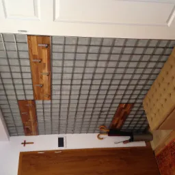 Metalowa krata na ścianie z drewnianymi panelami i haczykami, uzupełniona o wieszak na parasole, pufę i dywan w przedpokoju.