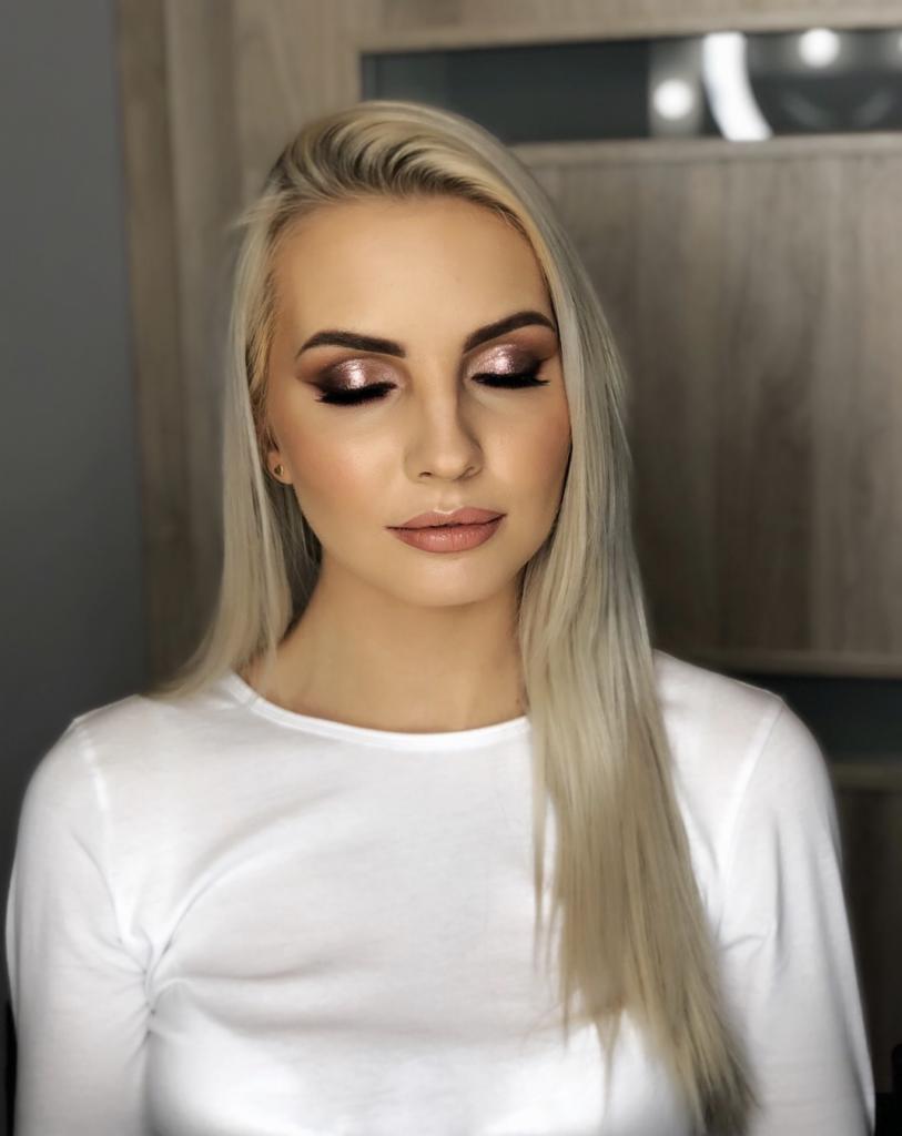 Portret kobiety z platynowymi włosami i profesjonalnym makijażem oczu w odcieniach brązu i złota, z zamkniętymi powiekami i delikatnym błyszczykiem na ustach, na neutralnym tle.