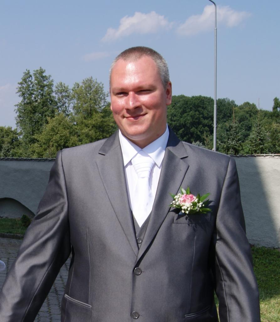 Uśmiechnięty mężczyzna w szarym garniturze z butonierką z różowych kwiatów na tle muru i zieleni.