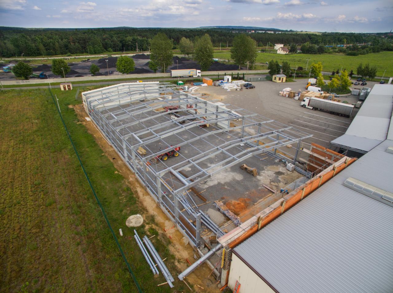 Szkielet stalowej konstrukcji hali w trakcie budowy, widok z góry na plac budowy z zaparkowanymi ciężarówkami i stertą węgla w tle.