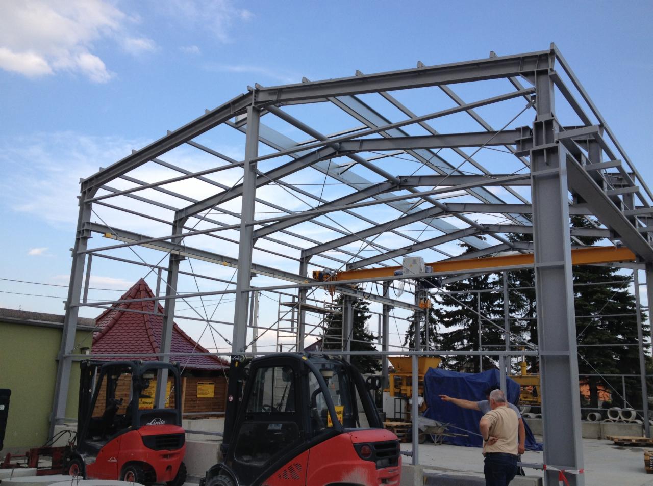 Stalowa konstrukcja hali w trakcie budowy, z widocznym suwnicą i dwoma wózkami widłowymi Linde na pierwszym planie, na tle błękitnego nieba.