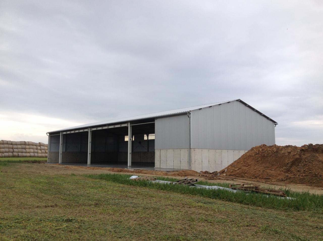 Konstrukcja stalowa hali magazynowej z szarej blachy trapezowej na fundamencie betonowym, widoczny stos bali słomy w tle i pryzma ziemi obok, pochmurne niebo.