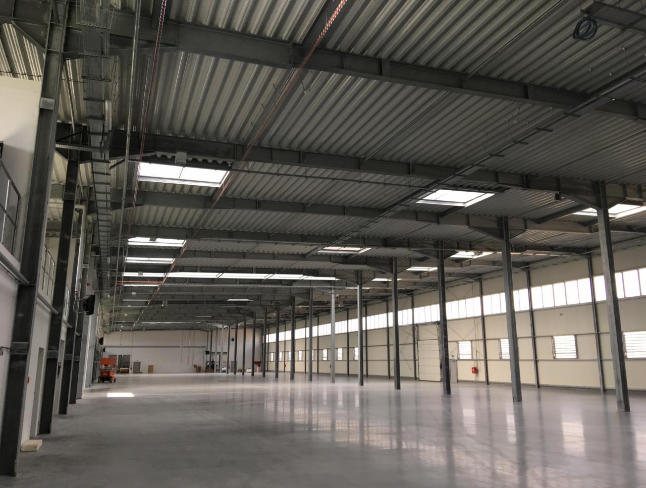 Wnętrze nowej, pustej hali przemysłowej z szarą, falistą blachą na dachu, metalowymi słupami i oknami w ścianach, jasna betonowa podłoga odbija światło, w oddali pomarańczowy podnośnik.