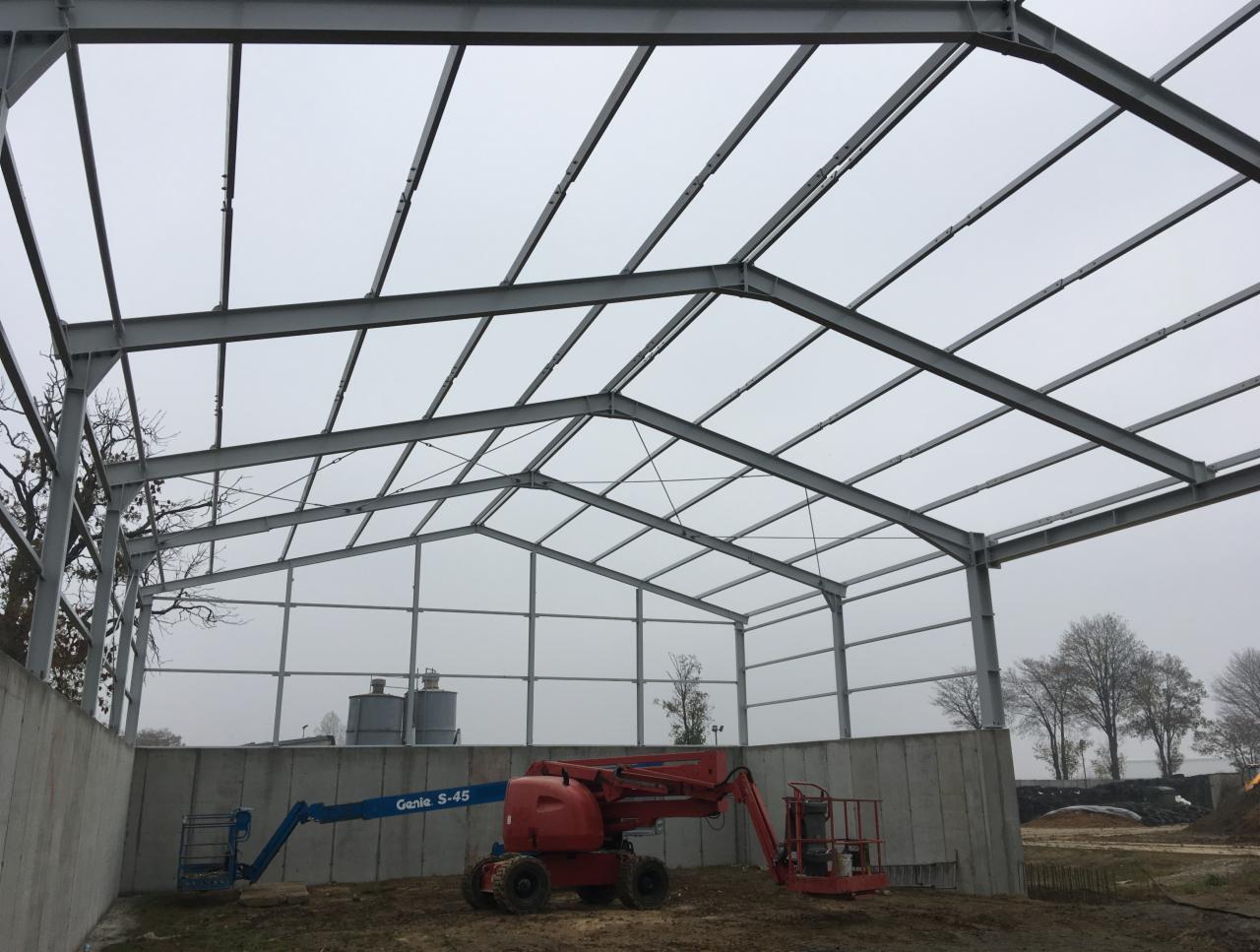 Montaż szkieletu stalowego hali z użyciem podnośników koszowych Genie S-45, widoczne silosy w tle.