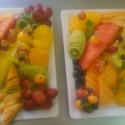 Dwa białe prostokątne talerze z kompozycjami z pokrojonych owoców: arbuz, melon, ananas, kiwi, pomarańcze, winogrona, physalis, udekorowane listkami mięty i małymi szpikulcami z owocami.