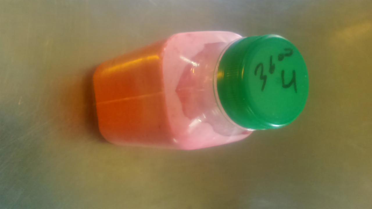 Plastikowa butelka z zieloną zakrętką, wypełniona różowo-pomarańczowym płynem, leżąca na metalowej powierzchni. Na zakrętce widoczny napis wykonany markerem.