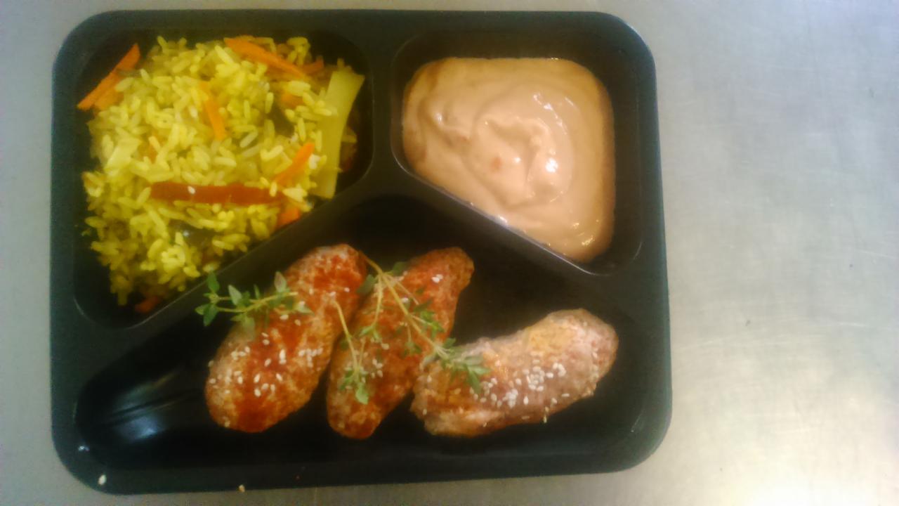 Czarna tacka z trzema przegródkami zawierająca ryż z warzywami, sos oraz trzy kawałki kurczaka posypane sezamem i udekorowane gałązkami tymianku.