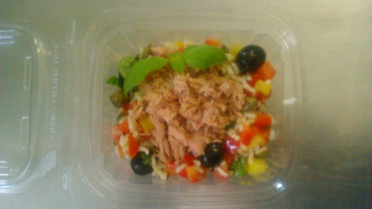 Sałatka ryżowa z tuńczykiem, papryką, oliwkami i kaparami, udekorowana listkami bazylii, podana w przezroczystym plastikowym pojemniku na wynos.