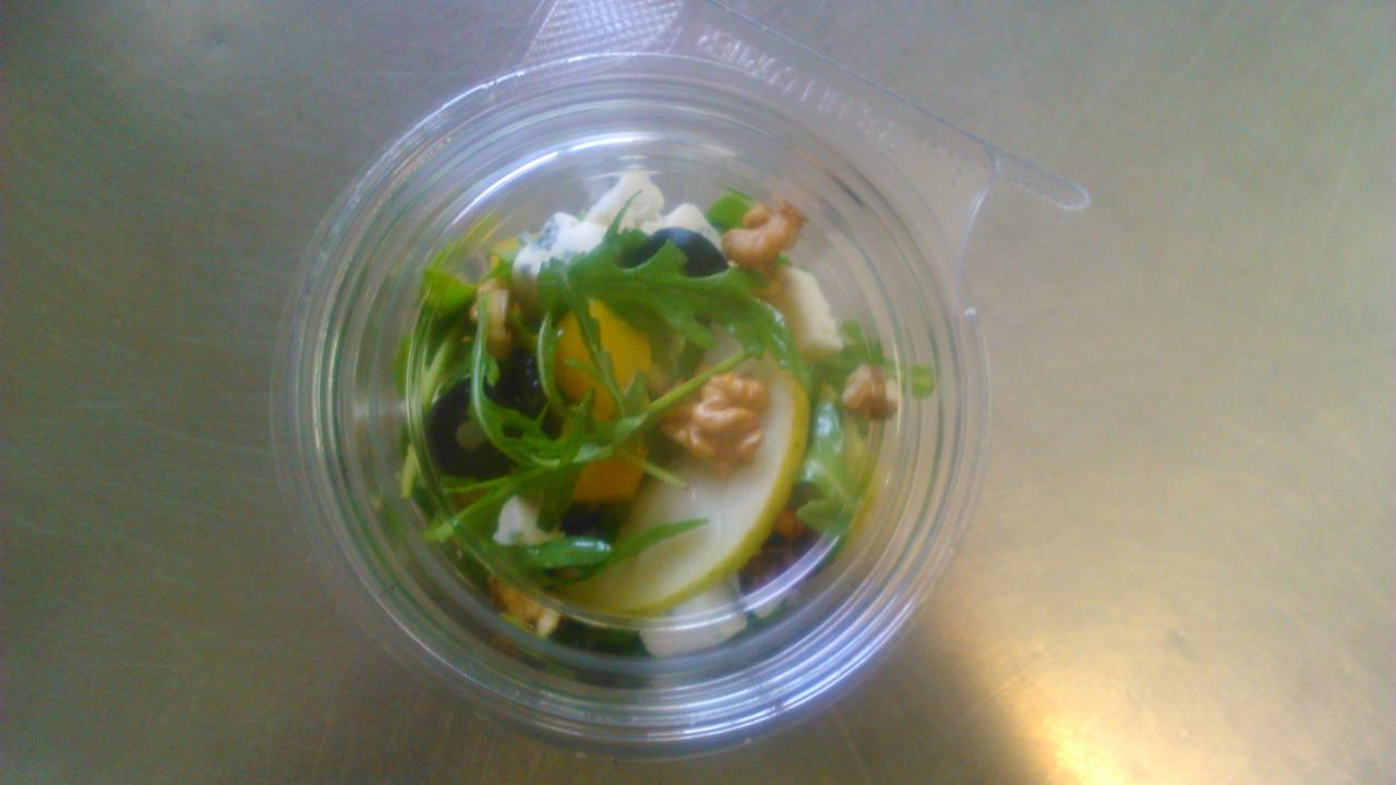 Sałatka z rukolą, gruszką, orzechami włoskimi, serem pleśniowym i pomarańczą w plastikowym opakowaniu na metalowym blacie.