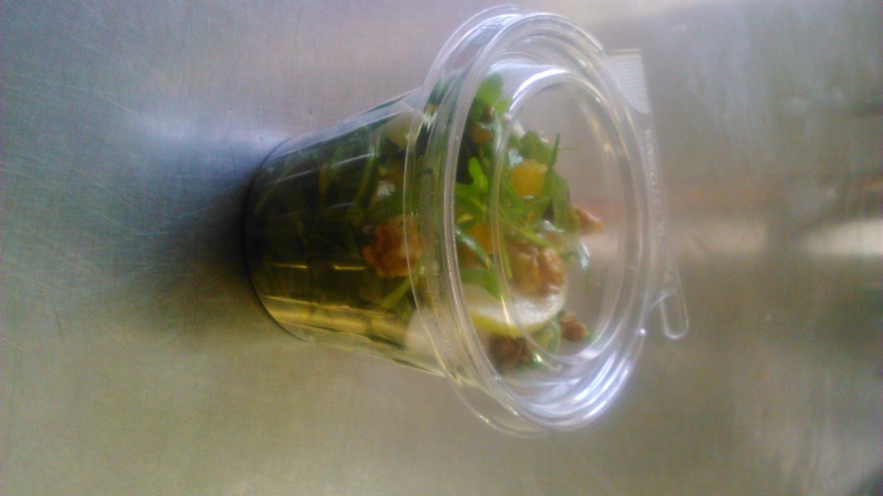 Sałatka z rukolą, orzechami i pomarańczą w przezroczystym plastikowym pojemniku na metalowym tle.