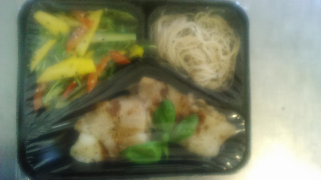 Zapakowany zestaw obiadowy w czarnym pojemniku: grillowana ryba z bazylią, sałatka z mango i warzywami oraz makaron ryżowy.