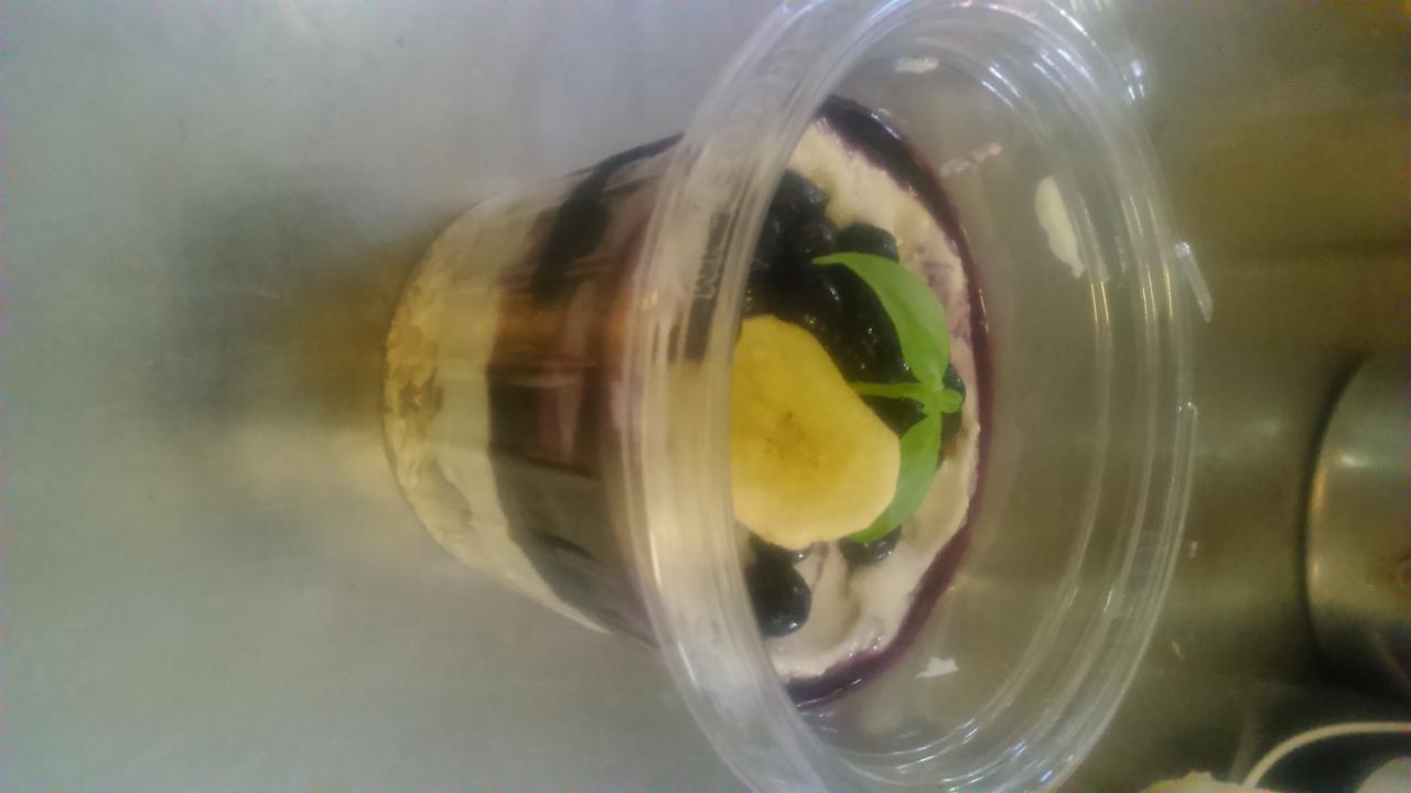 Deser warstwowy w przezroczystym pojemniku z widocznymi borówkami, bananem i listkiem bazylii na wierzchu, ułożony na metalowej powierzchni.