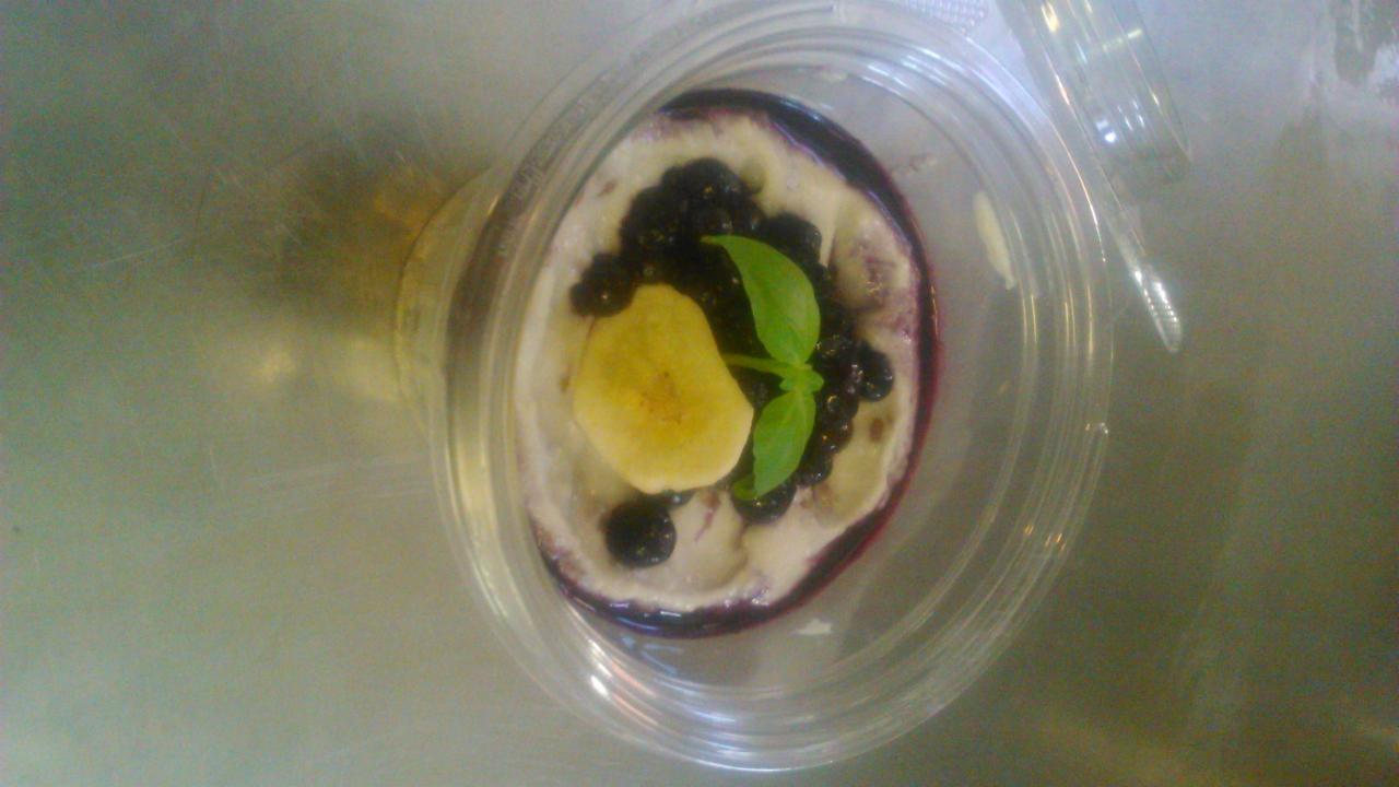 Deser w przezroczystym pojemniku z jogurtem, borówkami, bananem i listkami bazylii, widoczne fioletowe obrzeże.