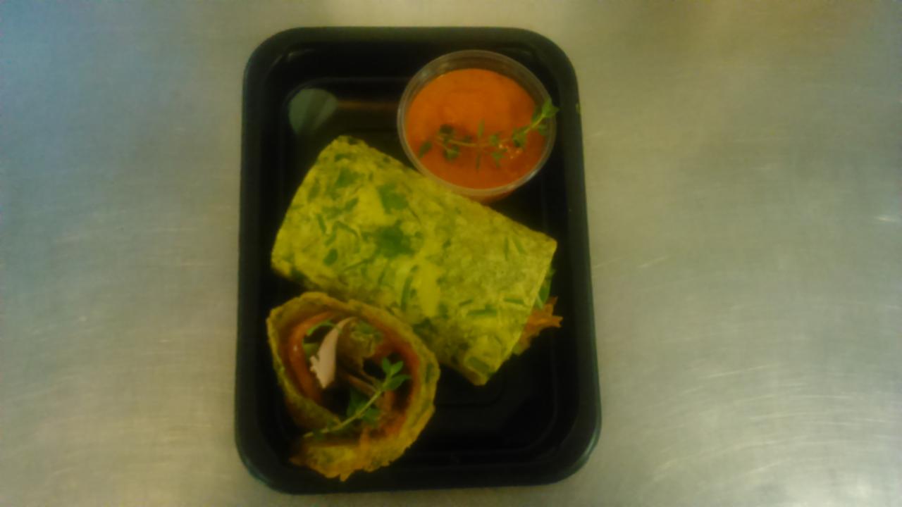 Wrap z zielonego ciasta z warzywami i mięsem, przekrojony na pół, podany z sosem pomidorowym i gałązką tymianku w czarnym pojemniku na tle metalowego blatu.