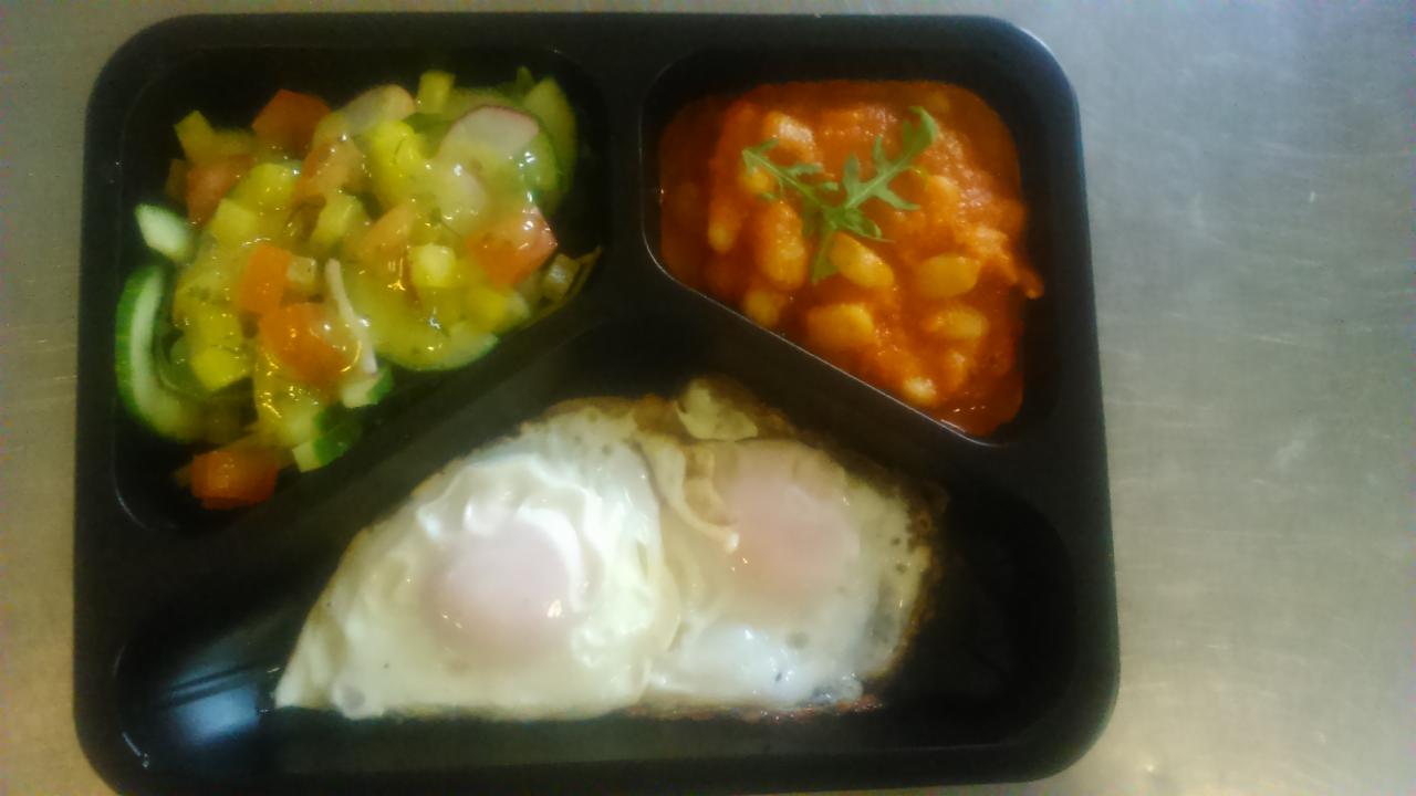 Zestaw obiadowy w czarnym pojemniku z trzema przegródkami: sałatka warzywna z ogórkiem i pomidorem, fasolka w sosie pomidorowym i dwa sadzone jajka.
