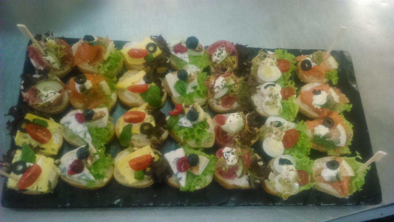 Różnorodne kanapki na ciemnej tacy, udekorowane sałatą, pomidorkami, jajkami, oliwkami, wędlinami, serami i owocami, niektóre z wykałaczkami.