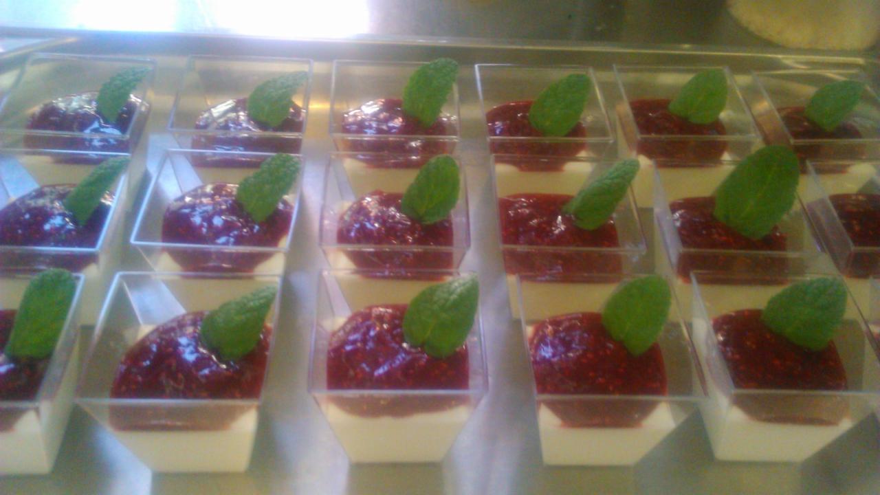 Deser panna cotta z sosem malinowym i listkiem mięty, podany w przezroczystych, kwadratowych naczyniach, ustawionych w rzędach na tacy.