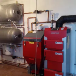 Kotłownia z dwoma szarymi zbiornikami akumulacyjnymi, czerwonym kotłem Defro i miedzianą instalacją hydrauliczną.