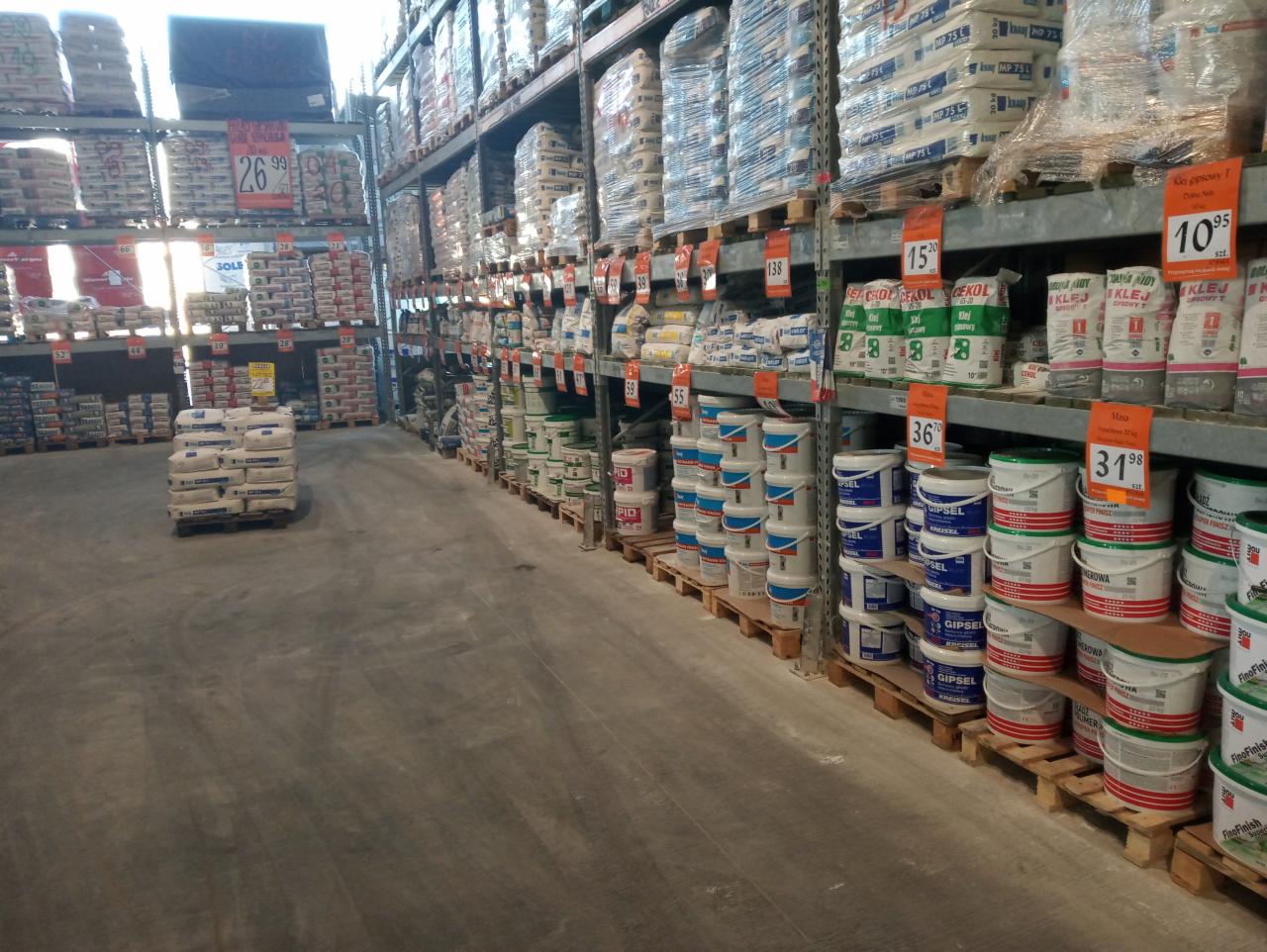 Wnętrze magazynu budowlanego z regałami wypełnionymi workami z zaprawami, klejami i farbami w wiaderkach, ustawionymi na paletach, z widocznymi cenami na etykietach.