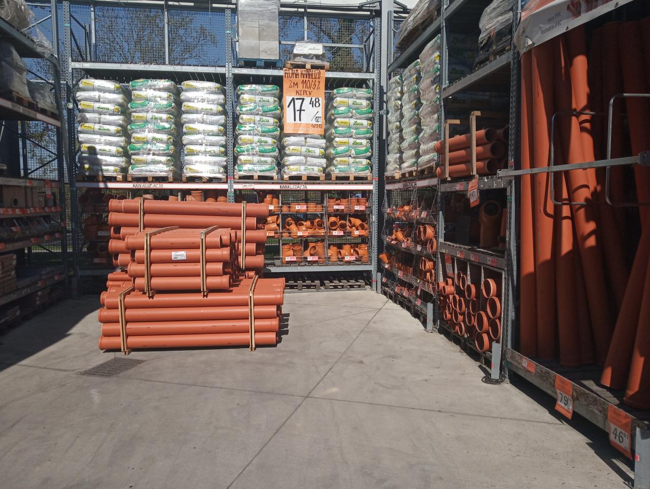 Wnętrze sklepu budowlanego z rzędami regałów wypełnionych workami z ziemią, elementami kanalizacji zewnętrznej i rurami PCV w odcieniach brązu i pomarańczy, ułożonymi na paletach i półkach.