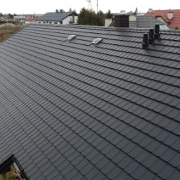 Nowy, grafitowy dach z dwoma oknami dachowymi i trzema kominami wentylacyjnymi na tle budującego się domu i krajobrazu.