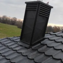 Czarny, metalowy komin wentylacyjny z poziomymi szczelinami, zamontowany na ciemnym dachu z falistej blachy na tle zielonego wzgórza i pochmurnego nieba.