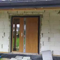 Montaż okien i drzwi wejściowych w surowym budynku z białych bloczków w Mielcu, widoczne drewniane podpory i niedokończone obróbki murarskie.