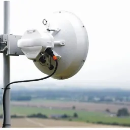 Biała antena telekomunikacyjna, zamontowana na metalowym maszcie, z widocznym kablem i uchwytami montażowymi, na tle rozmytego krajobrazu.