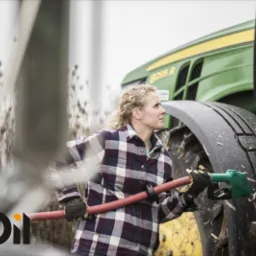 Kobieta w flanelowej koszuli tankuje zielony traktor John Deere za pomocą zielonego pistoletu do tankowania z czerwoną rurą, widoczne błoto na oponie.