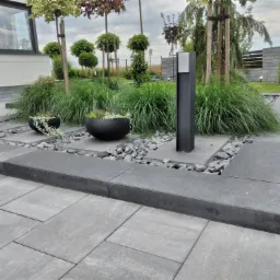 Ułożona szara kostka brukowa na posesji, z czarnymi donicami, ozdobnymi kamieniami i oświetleniem ogrodowym, w tle zadbana zieleń.