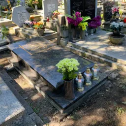 Granitowy pomnik nagrobny z ciemnego kamienia z wygrawerowanym krzyżem, ozdobiony bukietem jasnożółtych chryzantem i trzema lampionami, otoczony innymi nagrobkami na cmentarzu w słoneczny dzień.